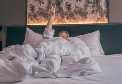 Nainen hotellin sängyssä peiton alla, näyttää sormilla peace-merkkiä