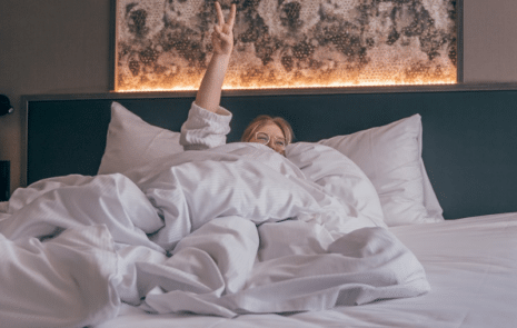 Nainen hotellin sängyssä peiton alla, näyttää sormilla peace-merkkiä
